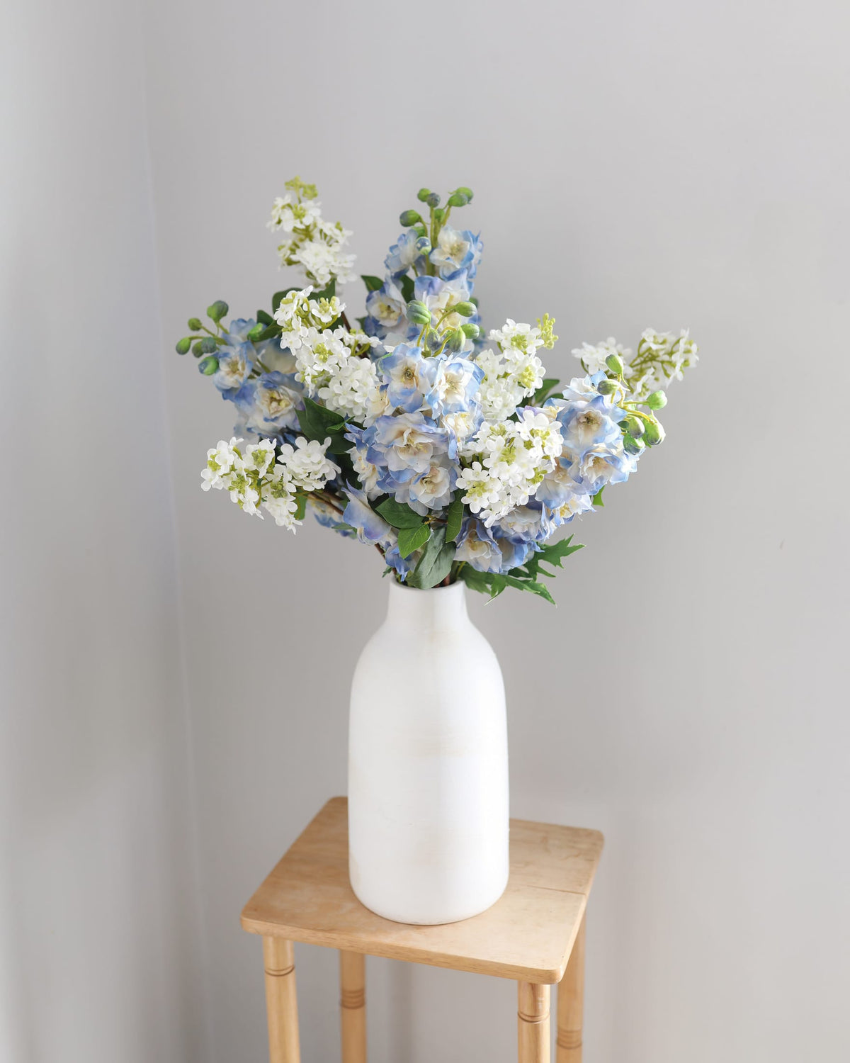 A bouquet of artificial blue and white delphinium and lilac arranged in a matte white ceramic vase, placed on a small wooden stool against a plain light gray wall.
