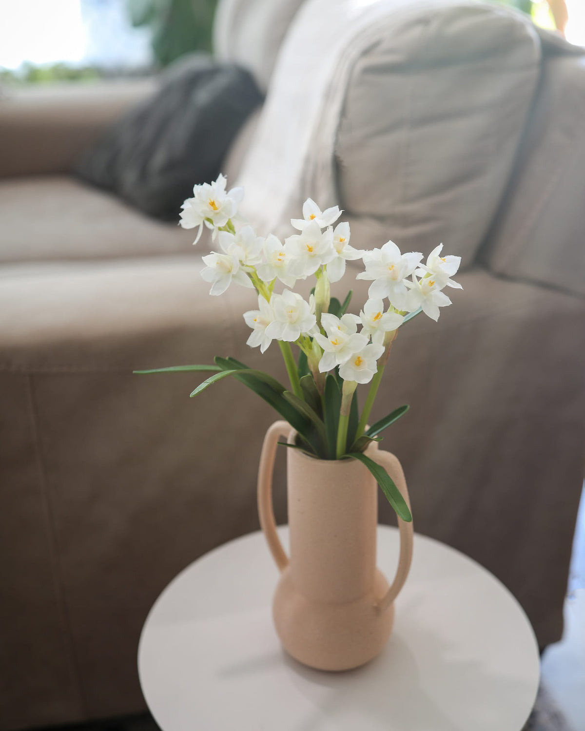 White faux Narcissus arranged in a beige ceramic pitcher vase, placed on a small round white side table in front of a blurred beige sofa.