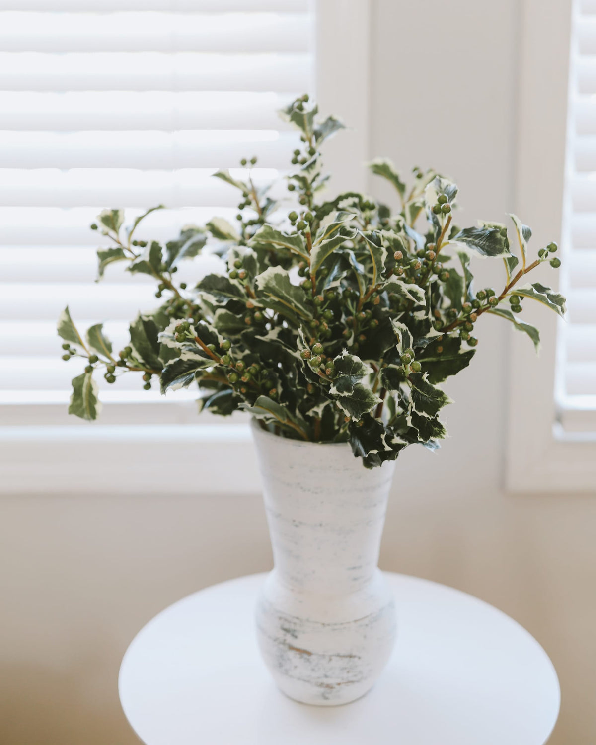 A decorative arrangement of green holly leaves and berries in a distressed white ceramic vase, set on a round white table in front of a softly lit window with white blinds.