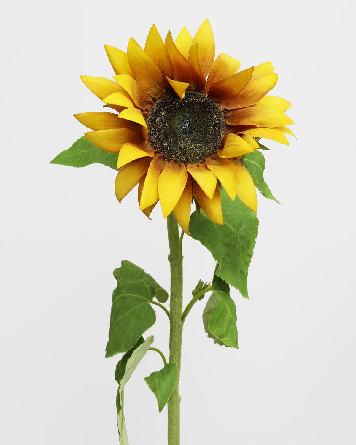 A single artificial sunflower with bright yellow petals, a dark brown center, and green leaves on a stem, photographed against a plain light gray background.