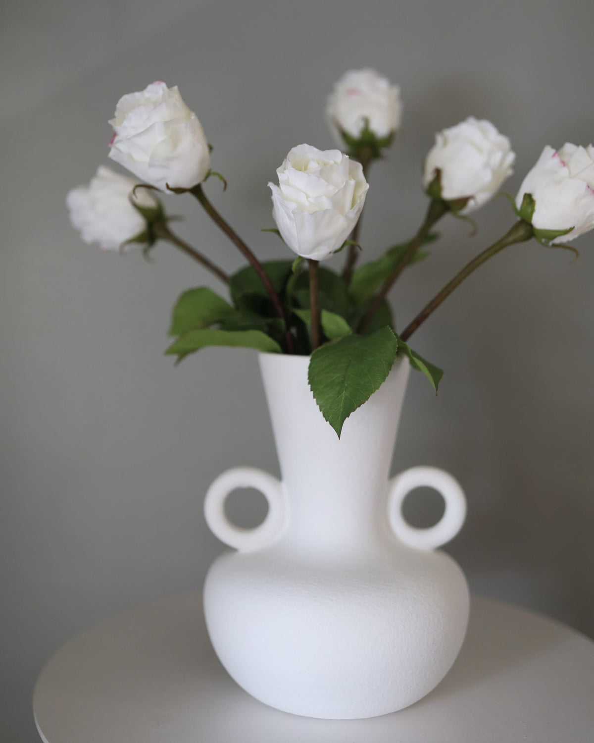White vase with two circular handles holding white faux roses on a gray background