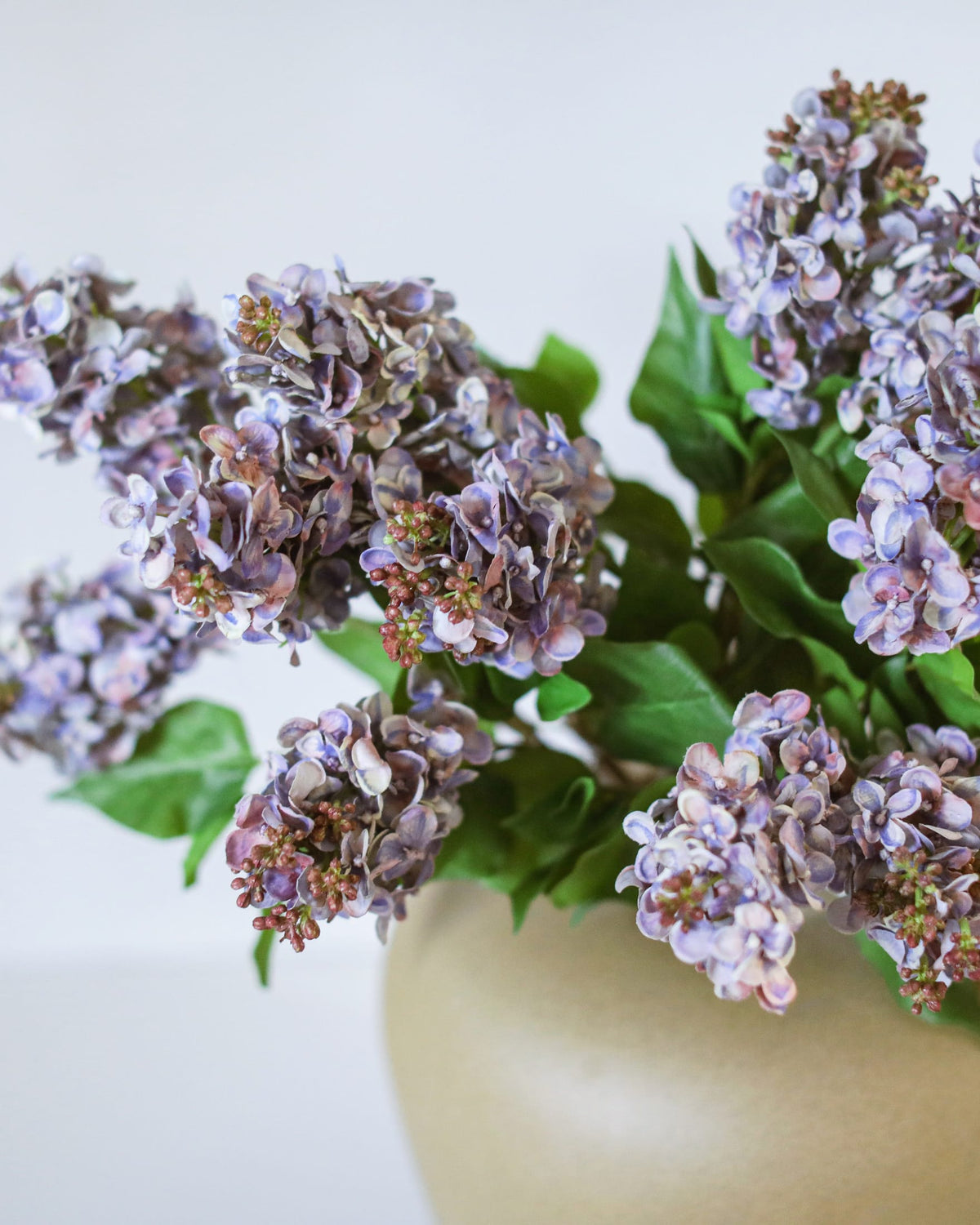 A close-up of artificial purple lilac flower clusters with green leaves arranged in a matte beige ceramic vase against a plain light background.