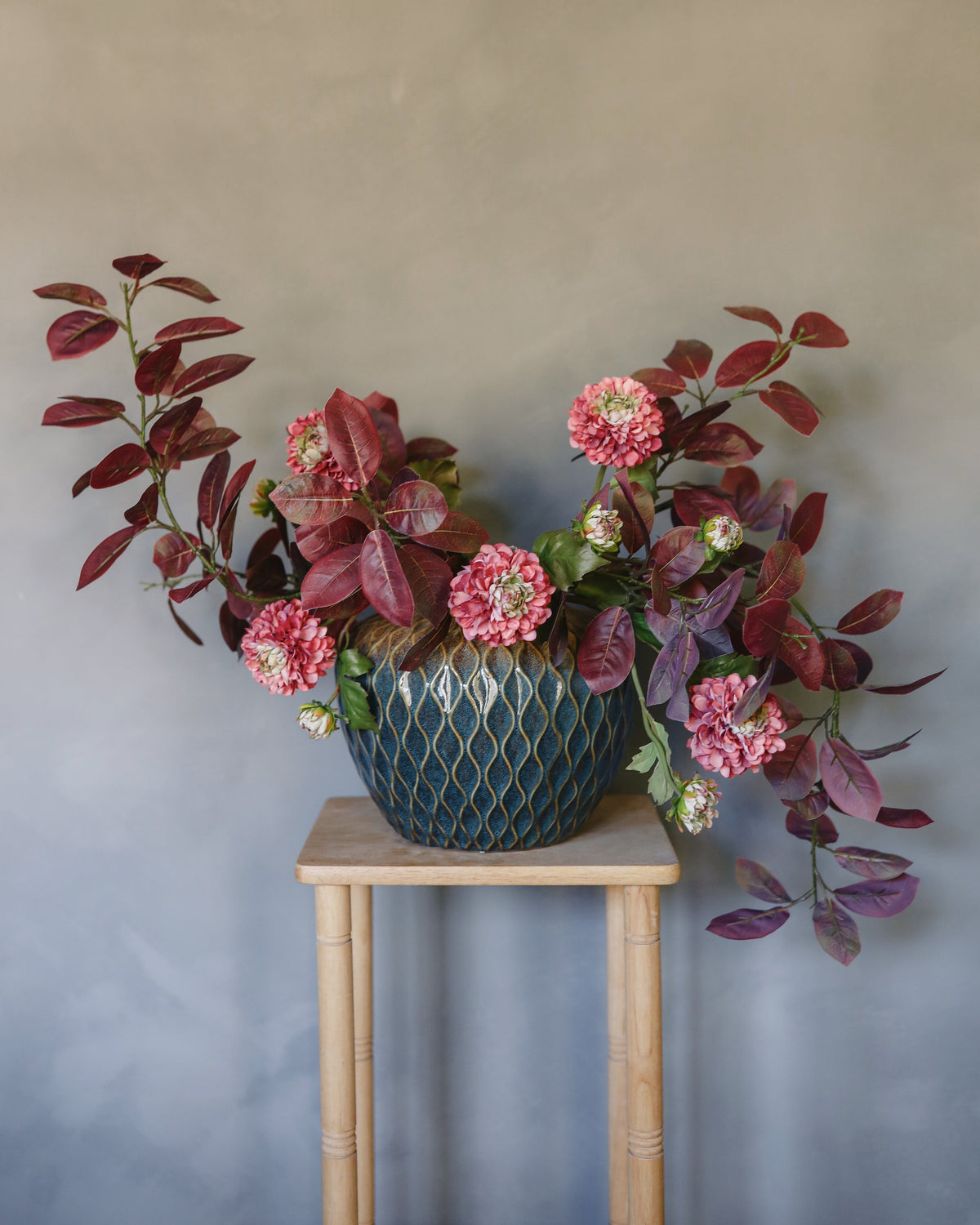 Floral smoketree and dahlia arrangement in a textured blue pot on a wooden stool against a plain wall.