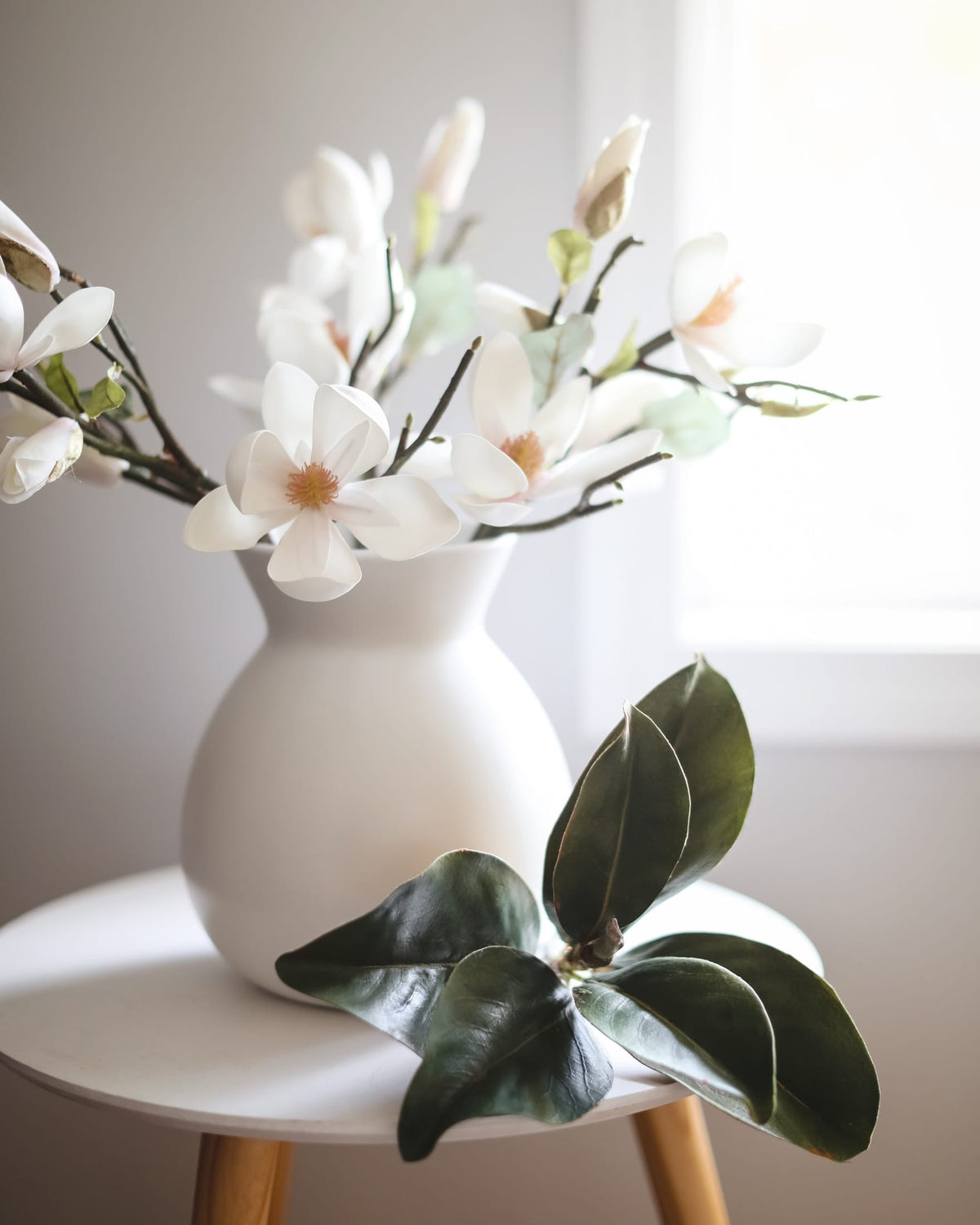 White ceramic vase filled with faux white magnolia branches, styled on a white side table with a green leafy stem in the foreground against a soft neutral background.