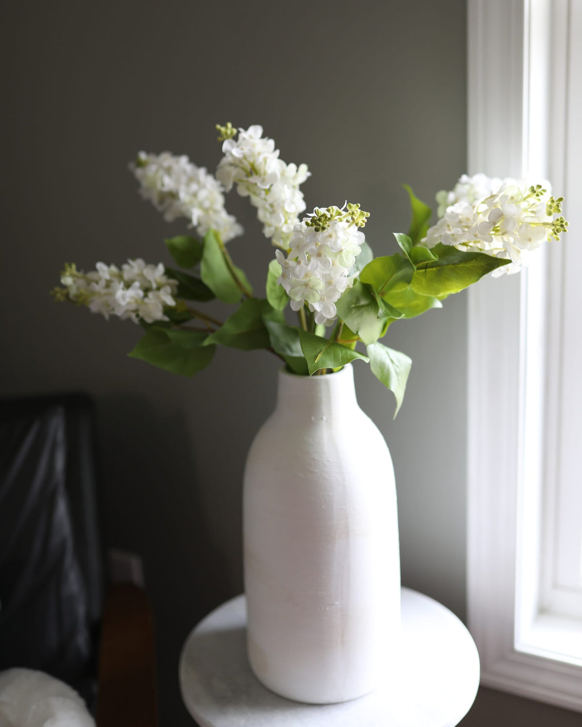 White faux lilac artificial flowers with green leaves arranged in a textured white ceramic vase, set on a round white side table in a dimly lit room.