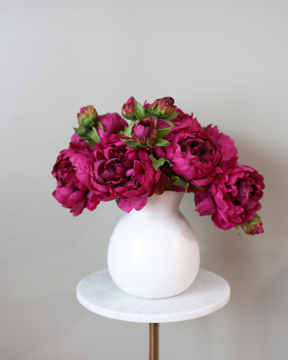 A bouquet of faux deep magenta peonies with green leaves and buds, arranged in a matte white ceramic vase, displayed on a small round white marble side table with a gold metal stand leg against a neutral light gray background.