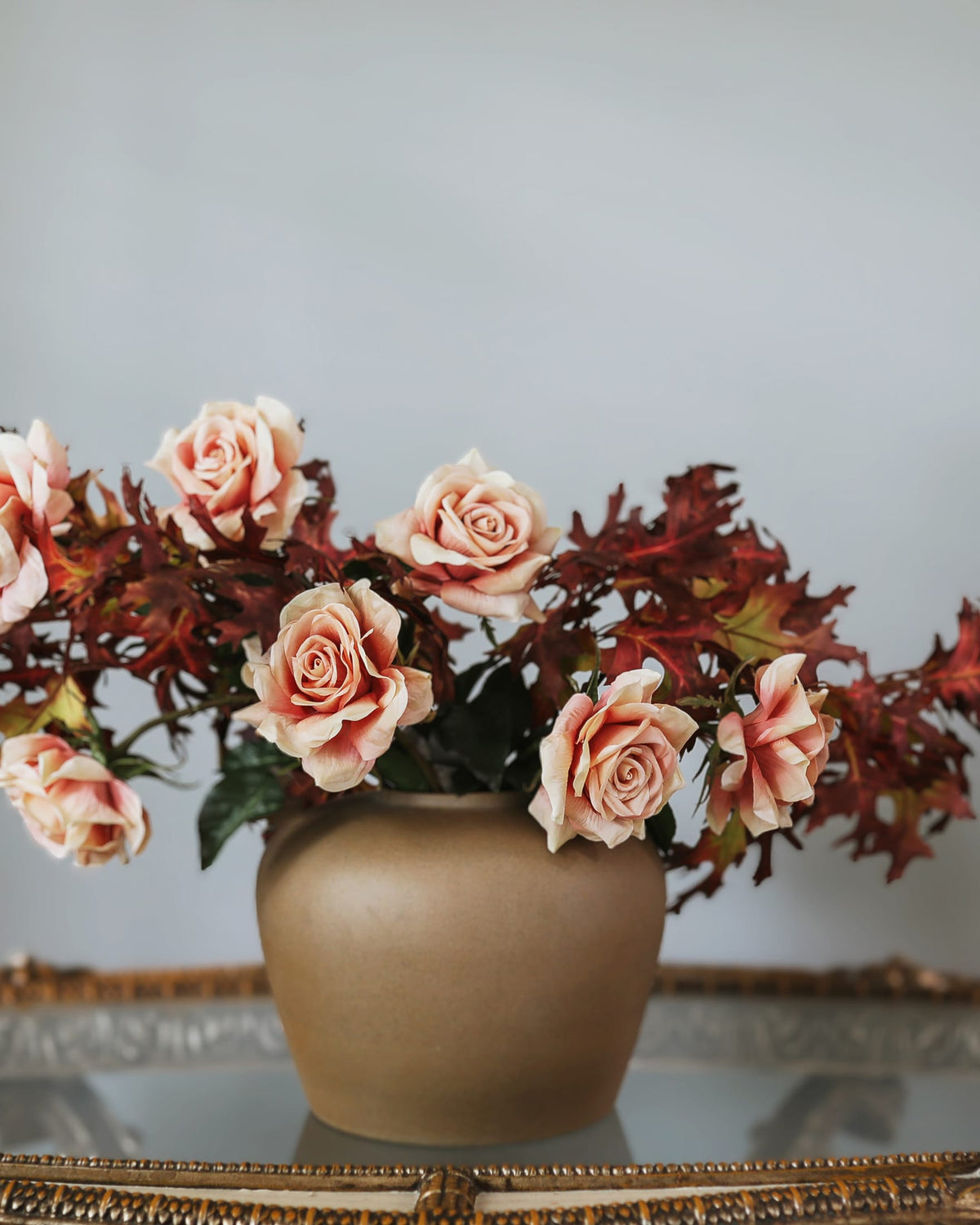 A decorative arrangement of artificial roses in soft blush pink and deep burgundy-red tones, displayed in a matte taupe ceramic pot on a reflective surface against a light gray background.