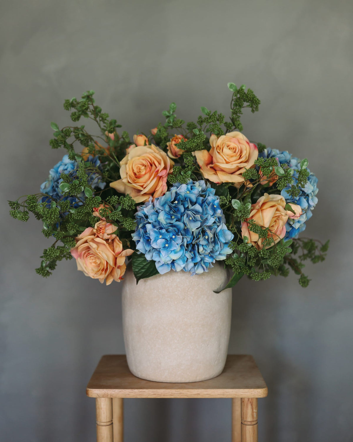 A faux loral arrangement of peach roses, blue hydrangeas, and green foliage in a textured cream ceramic pot, displayed on a small wooden stool against a gray wall.