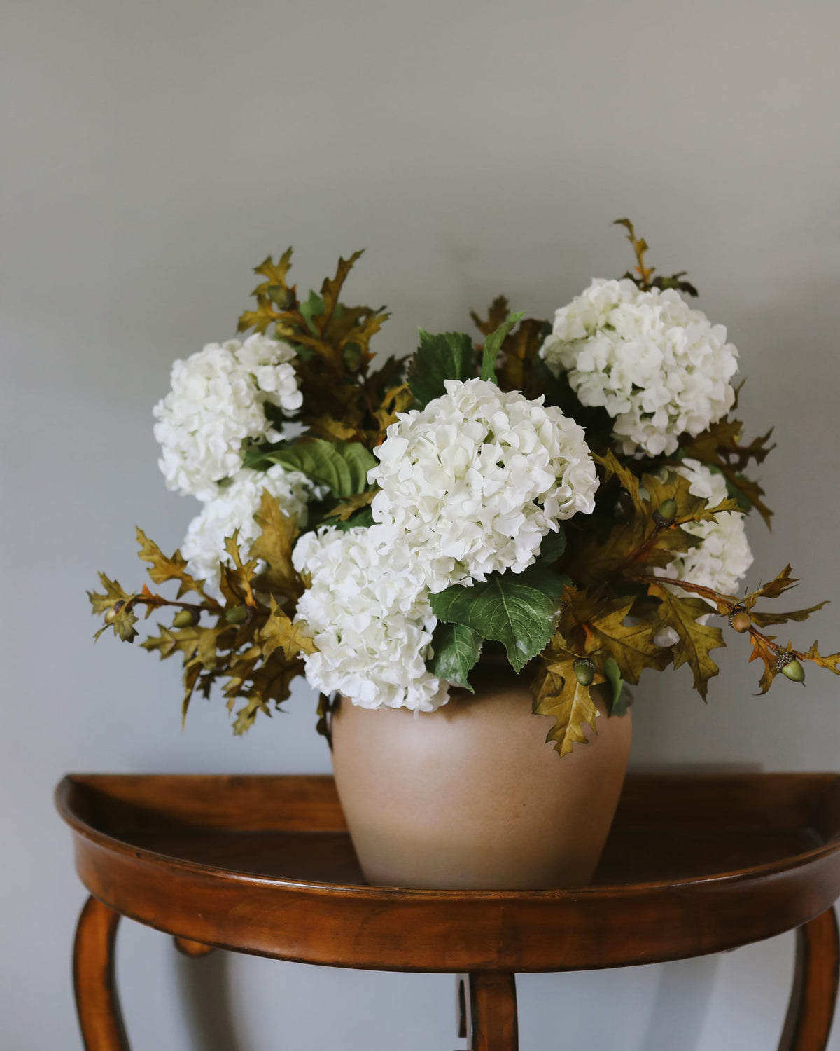 A floral arrangement of faux white hydrangea-like blooms with green and bronze-toned leaves in a matte beige pot, displayed on a wooden side table against a plain light gray wall.