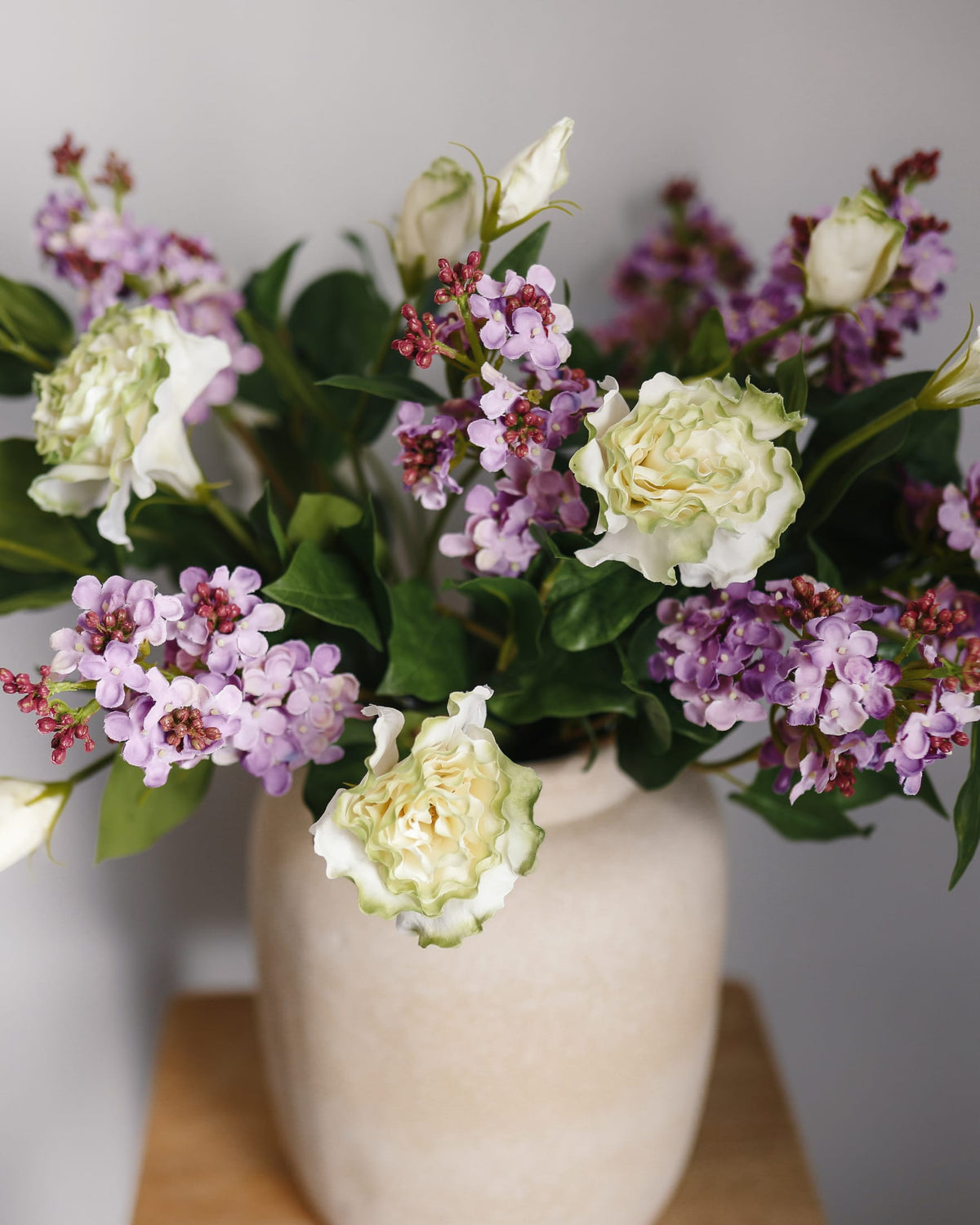 A bouquet of artificial flowers in a textured cream-colored ceramic vase, featuring white and pale yellow roses, purple lilac, and green leaves, set against a neutral gray background.