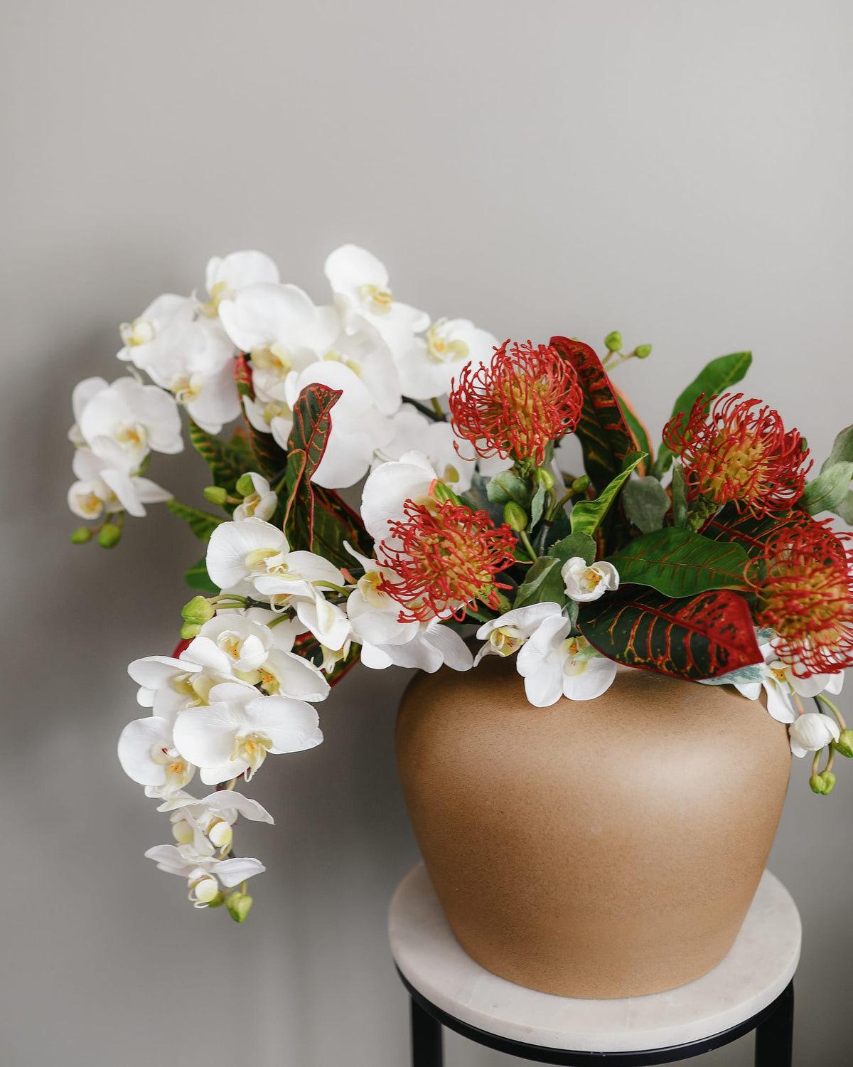 White artificial orchids with tropical proteas arranged in a bronze vase.