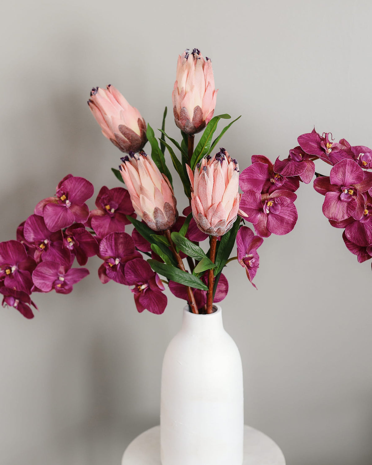 A floral arrangement featuring artificial pink protea-like flowers and deep magenta orchids with green leaves, displayed in a matte white ceramic vase against a soft light gray background.