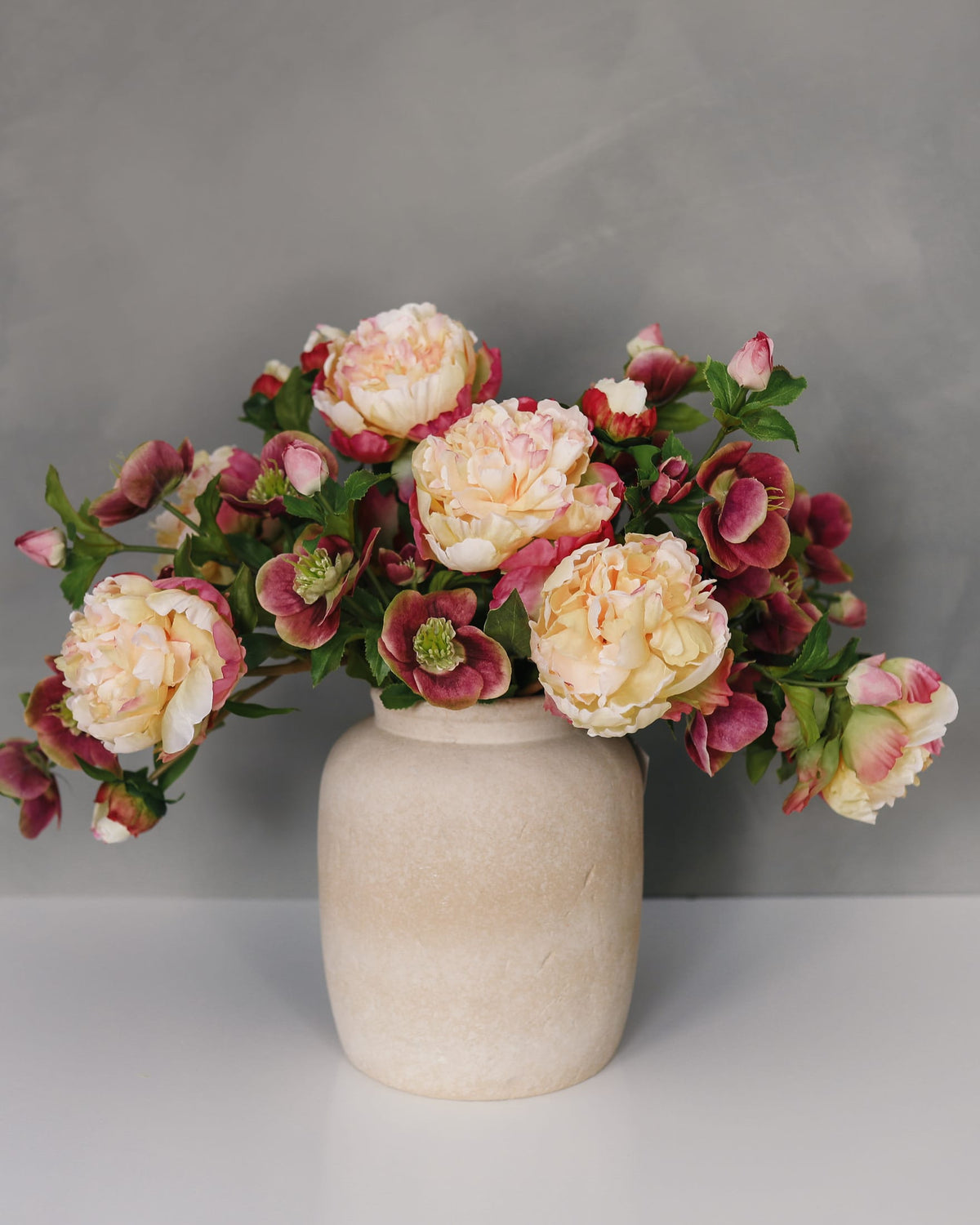 A bouquet of artificial peony and hollyhock-style flowers in soft cream, blush pink, and burgundy tones with green leaves, arranged in a matte beige ceramic vase against a gray studio background.