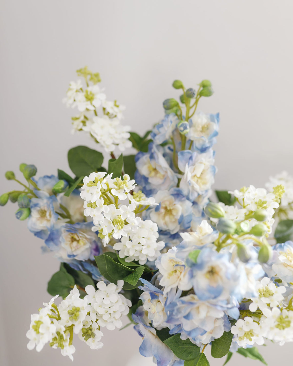 A bouquet of artificial flowers featuring light blue and white lialc with green leaves, arranged against a plain light gray background.