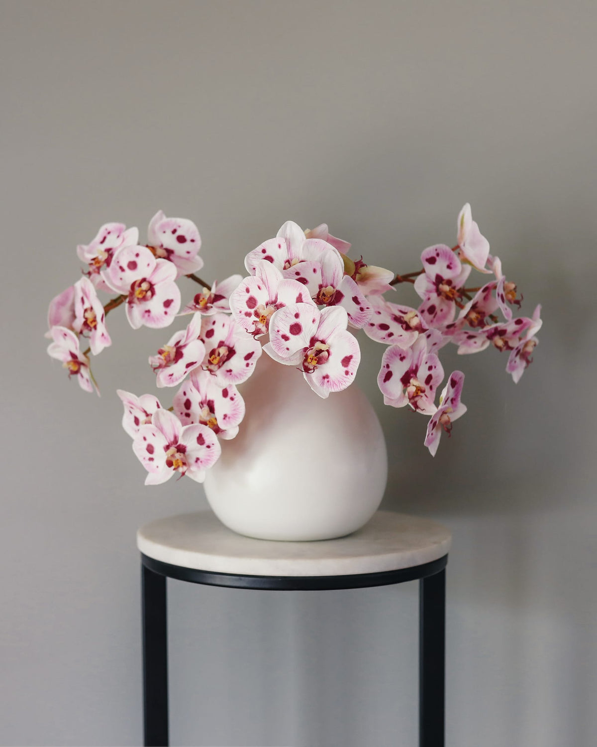 Pink and white artificial orchid flowers arranged in a matte white ceramic vase, displayed on a small round table with a light stone top and black metal frame against a neutral gray wall.