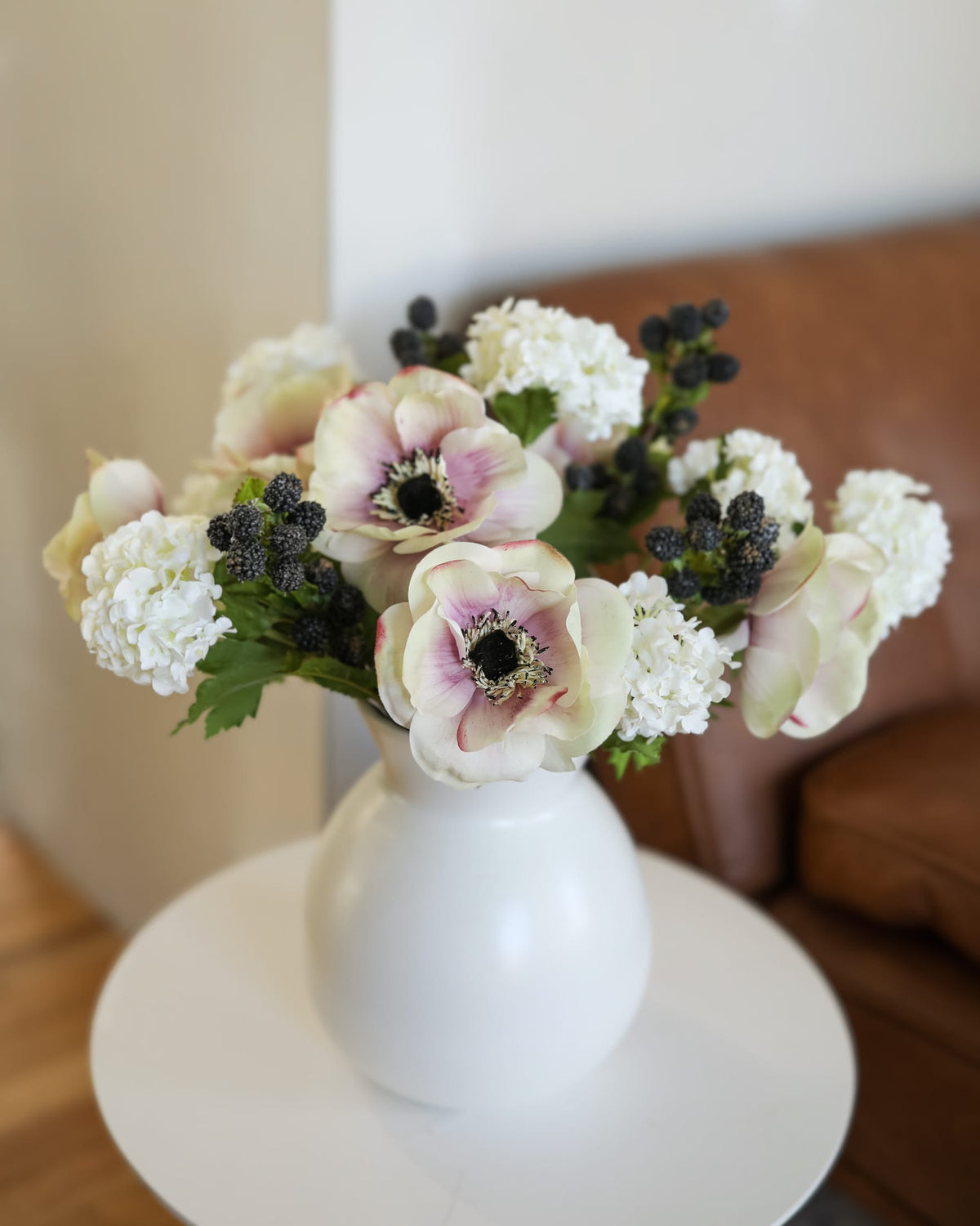 A white ceramic vase filled with a floral arrangement of artificial anemone flowers, white hydrangeas, and blackberries, displayed on a round white side table in a living room setting.