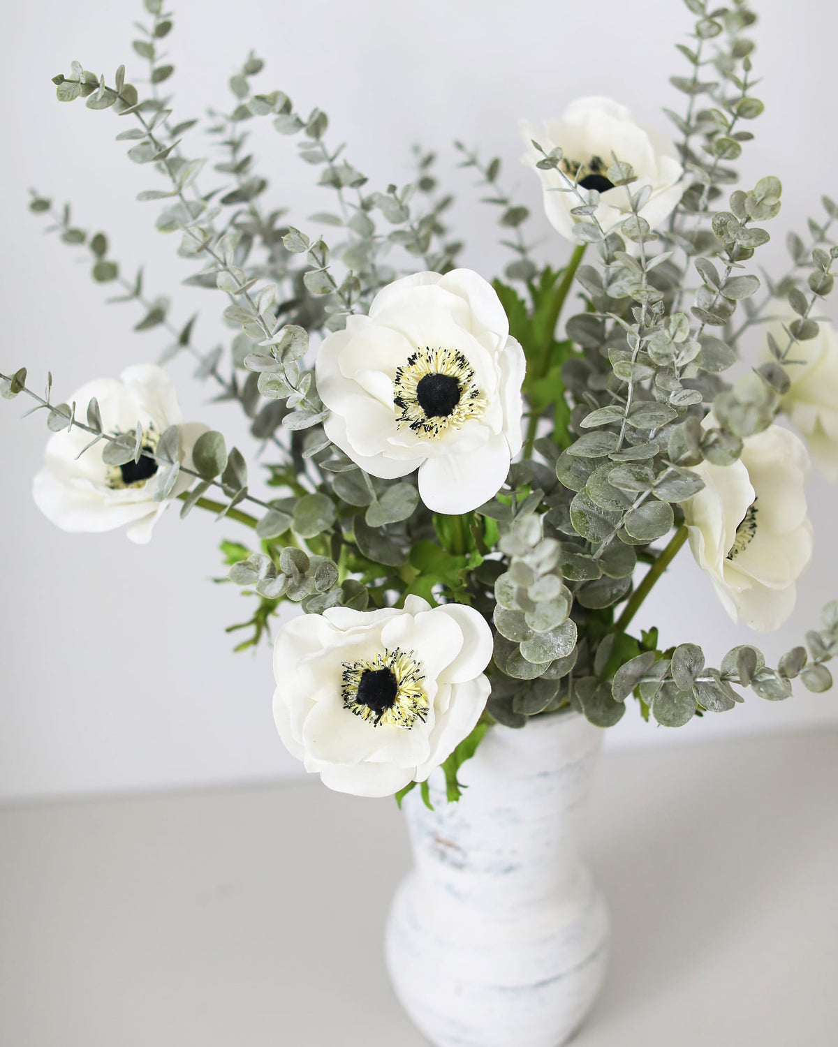 A bouquet of artificial white anemone flowers with dark centers and silver-green eucalyptus stems, arranged in a distressed white ceramic vase against a clean white background.