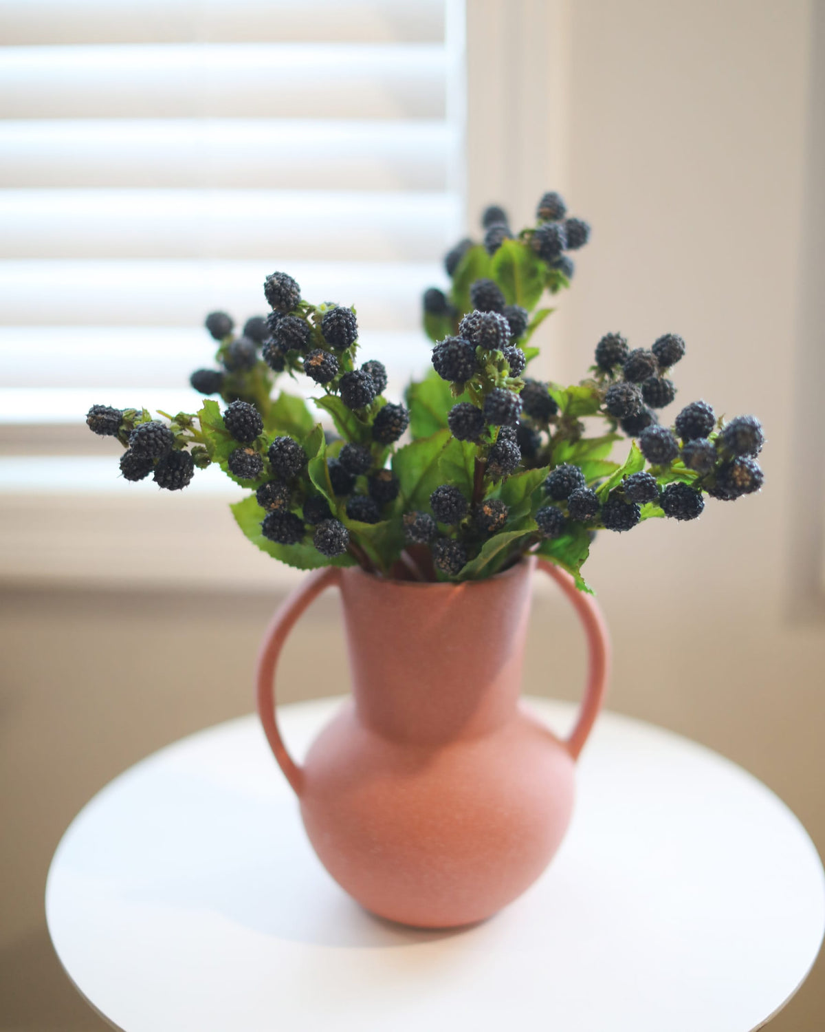 A terracotta-colored ceramic pitcher vase with two handles, filled with artificial blackberries and green leaves, displayed on a white round table in front of a window with white blinds.
