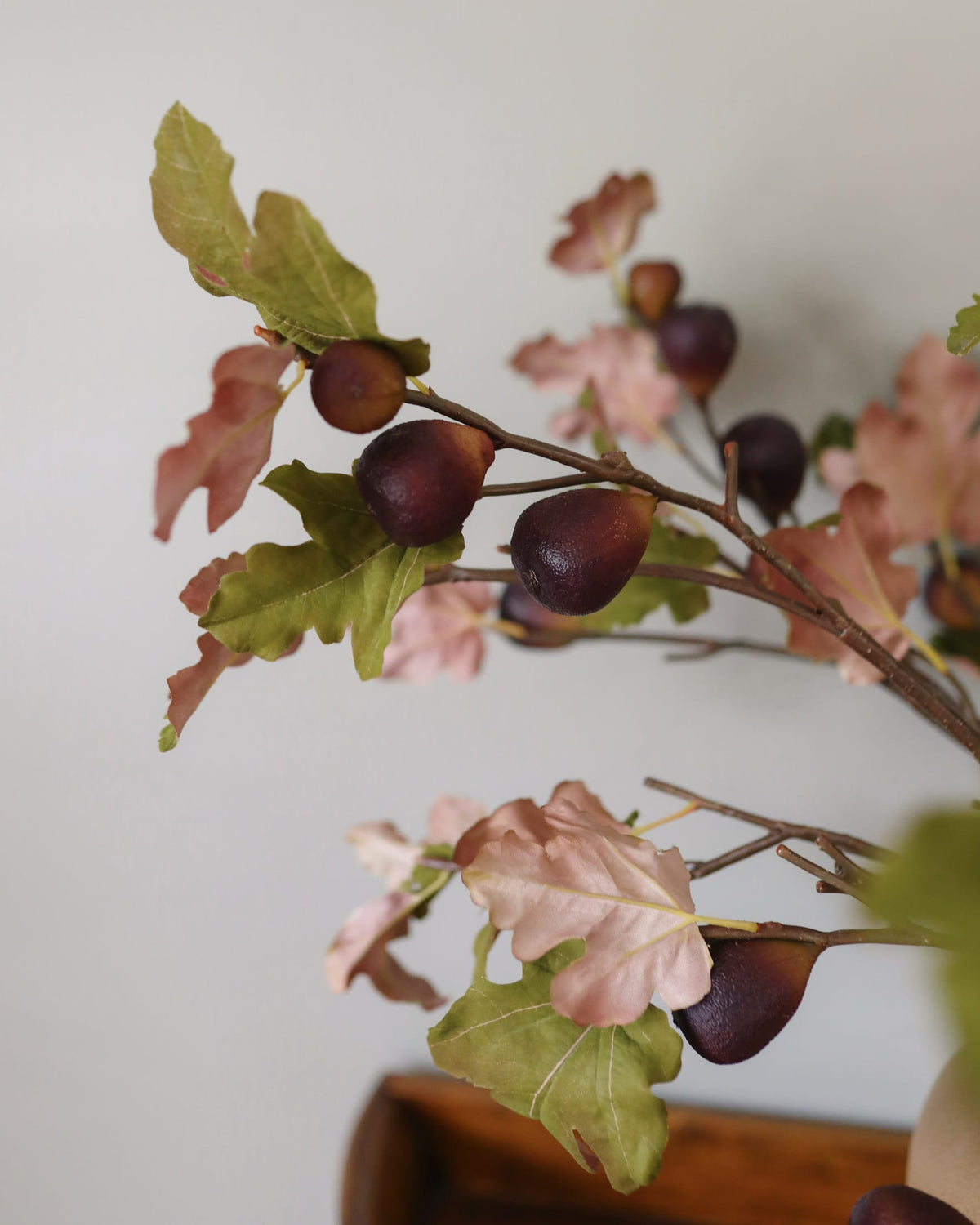 Artificial fig branch with realistic brown acorns, green and pinkish autumn leaves, and brown twigs, set against a plain light gray wall.