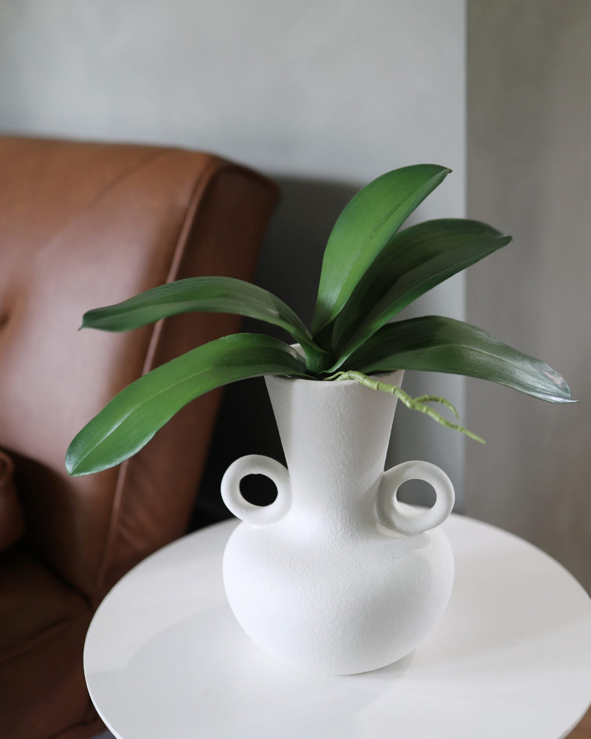 A white ceramic planter with two loop handles, holding a green faux orchid leaves set on a white round side table next to a brown leather armchair.
