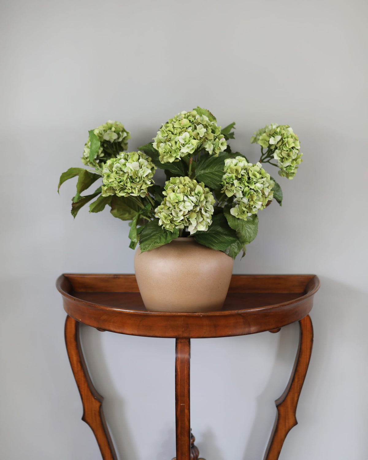 Vase with faux green hydrangeas on a wooden side table against a plain wall