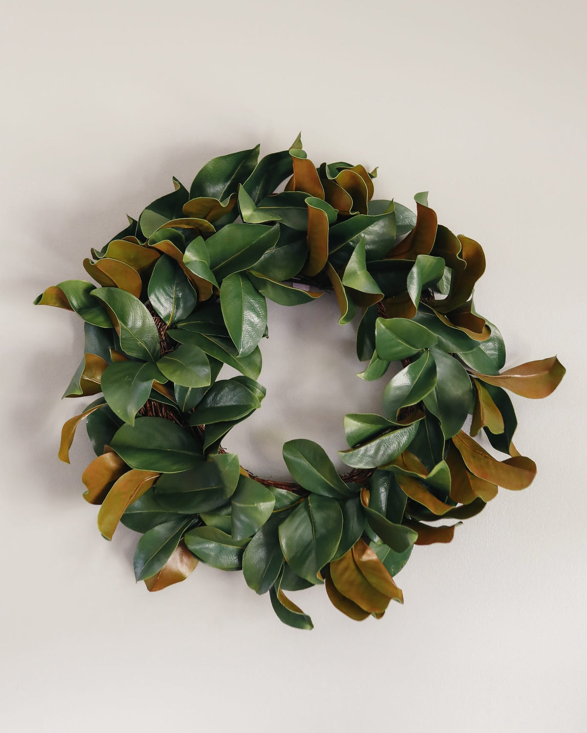 An artificial magnolia leaf wreath with glossy green and tan-brown leaves, photographed against a plain light gray background.
