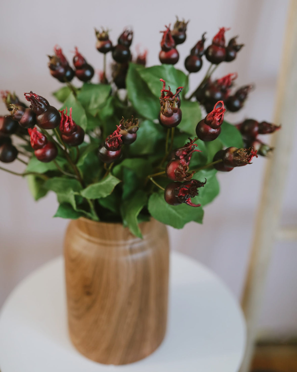 A bouquet of artificial rose hips arranged in a round wooden vase, placed on a white round table against a soft light background.