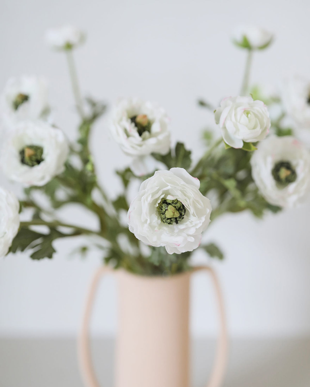 White faux ranunculus flowers with green centers arranged in a pale blush pink ceramic pitcher vase against a soft, light gray background.