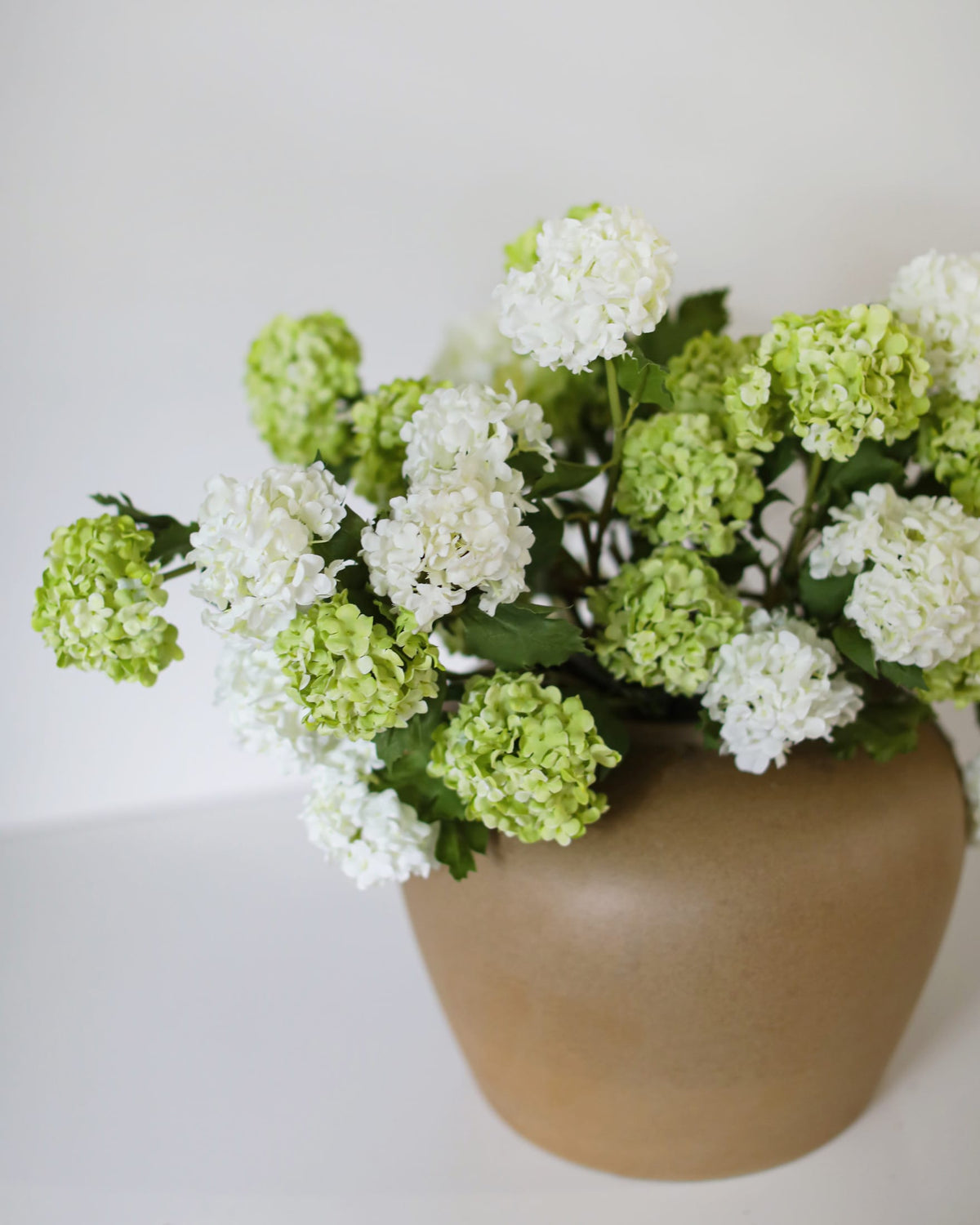 A bouquet of fake white and light green hydrangea-style snowballs flowers arranged in a matte beige ceramic vase against a clean white background.