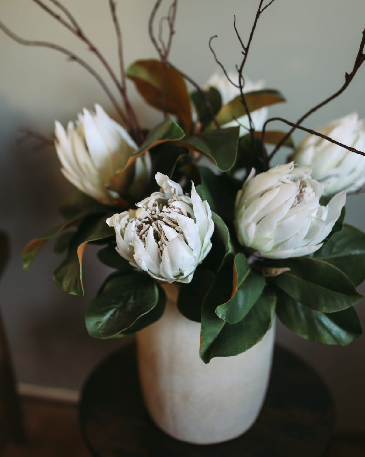Faux floral arrangement with white flowers and magnolia leaves in a white vase on a blurred background