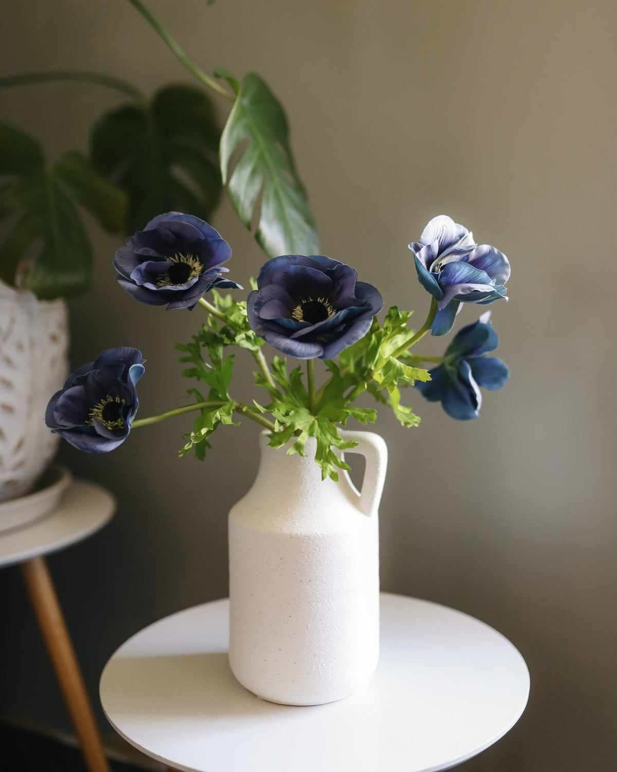 A white ceramic pitcher vase holding a bouquet of dark blue faux anemone flowers with green foliage, styled on a small round white side table against a neutral gray wall.