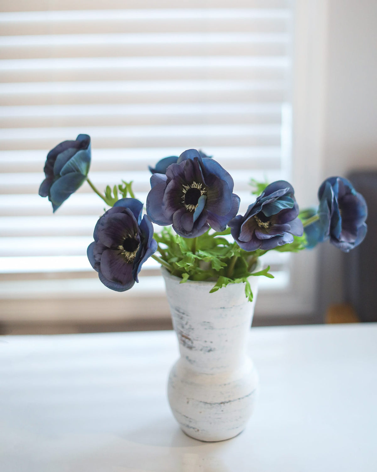 A bouquet of dark blue faux anemone flowers with green leaves arranged in a distressed white ceramic vase, set on a white table in front of a softly blurred window with white blinds.