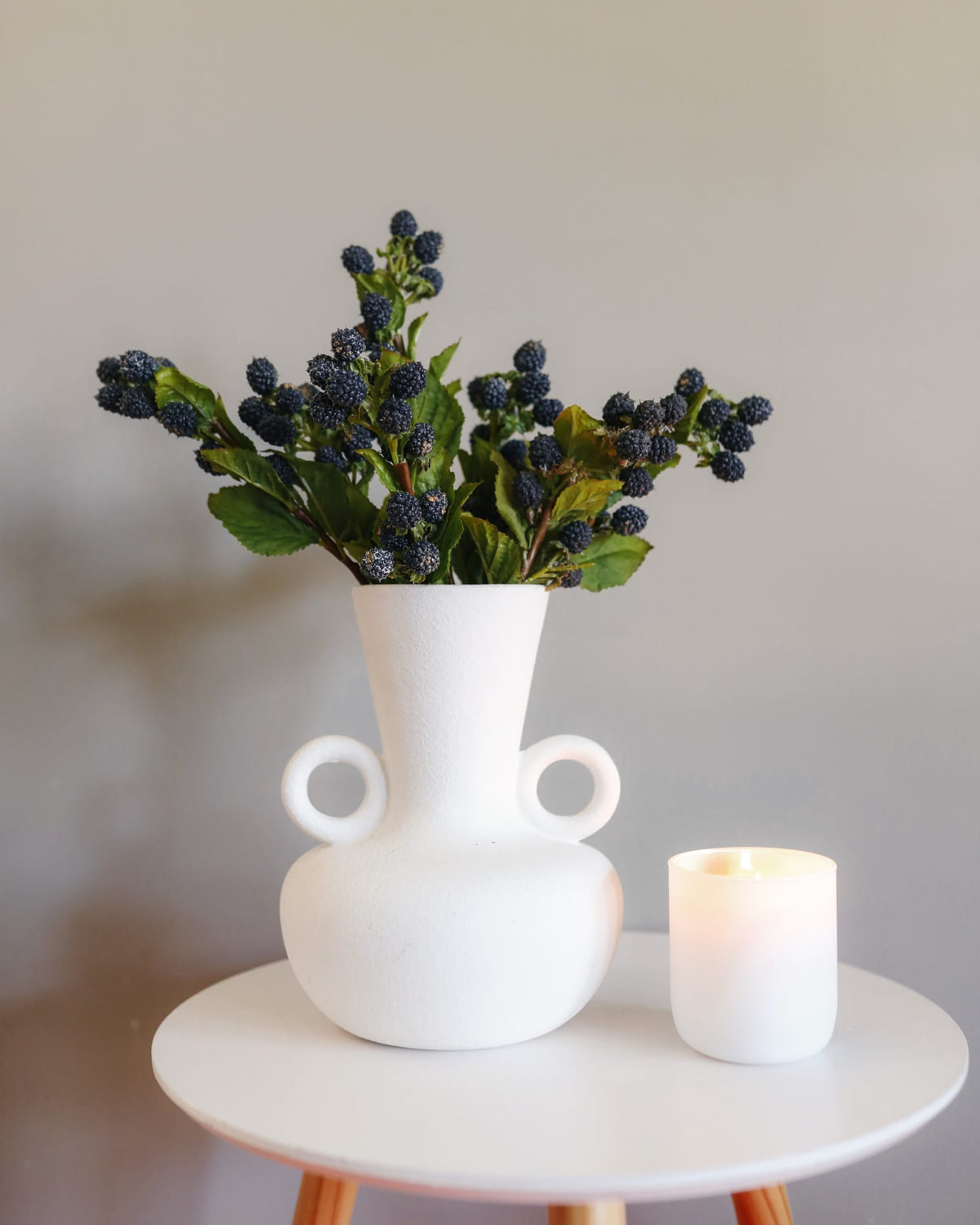 A matte white ceramic vase with two side loops, holding faux blackberries with green leaves, styled on a white round table next to a lit white candle.