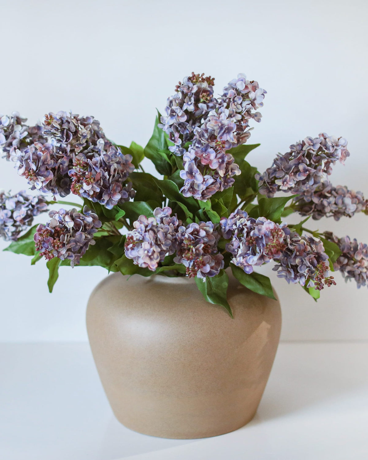 A faux purple-blue lilac arrangement in a beige ceramic vase against a plain white background.