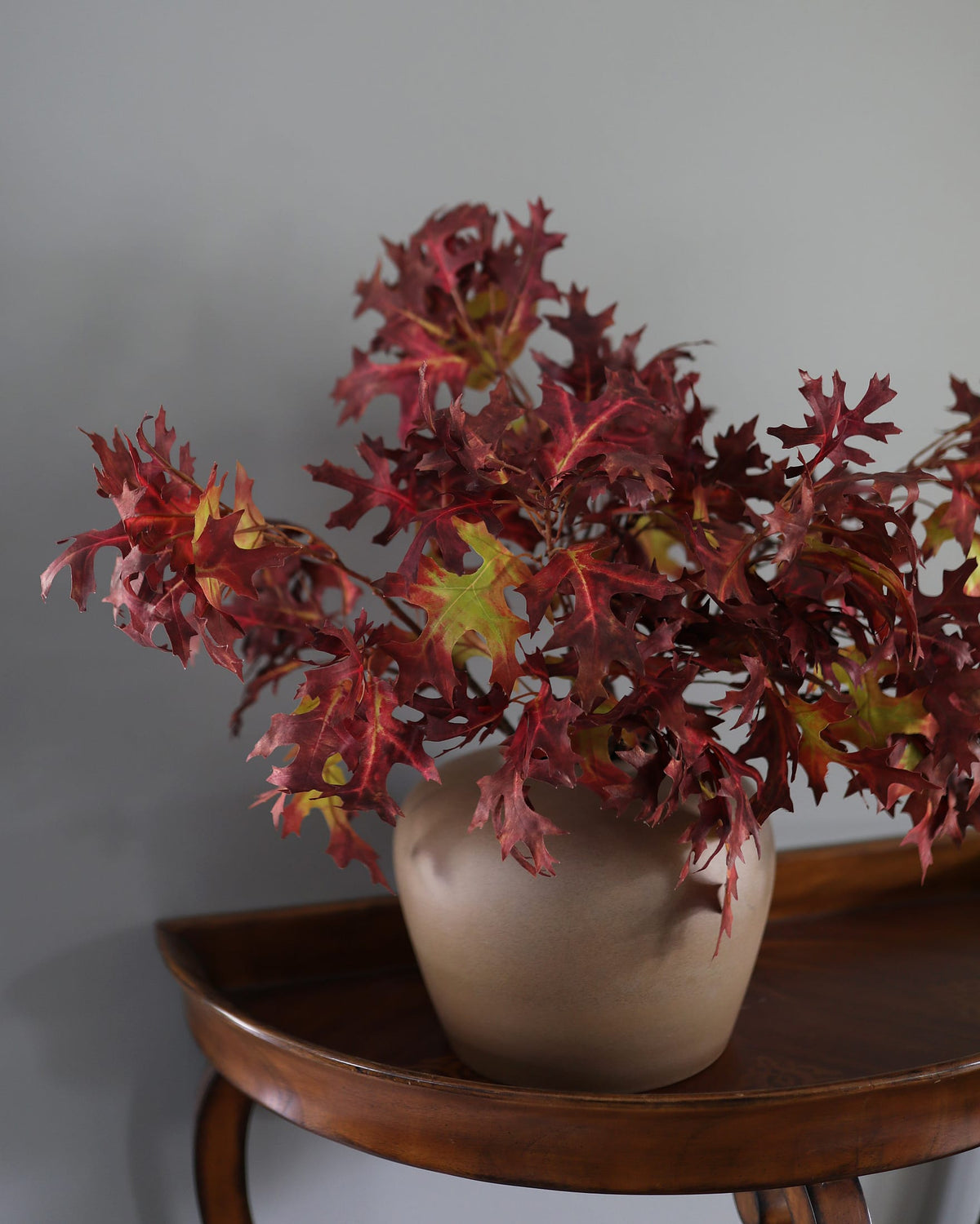 An artificial autumn leaf arrangement in deep red and burgundy tones with a few yellow-green accents, displayed in a matte beige ceramic pot on a dark wood side table against a neutral gray wall.