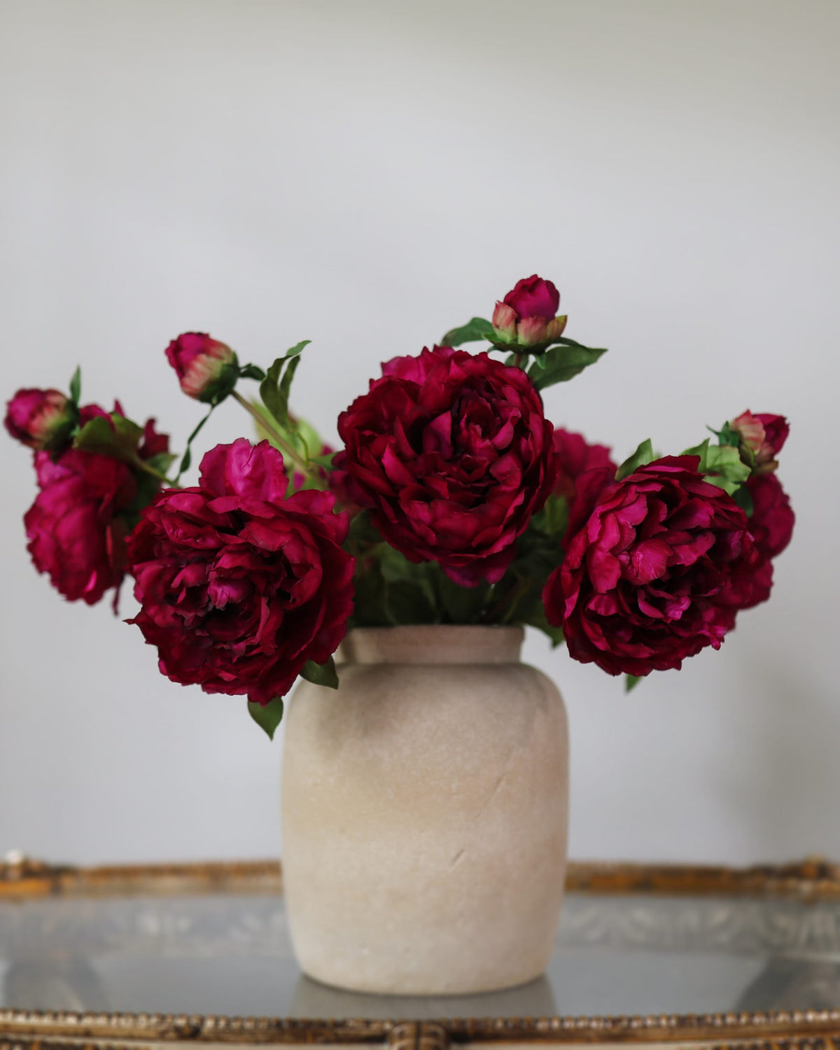 A bouquet of deep magenta faux peonies with green leaves and buds, arranged in a ceramic vase.