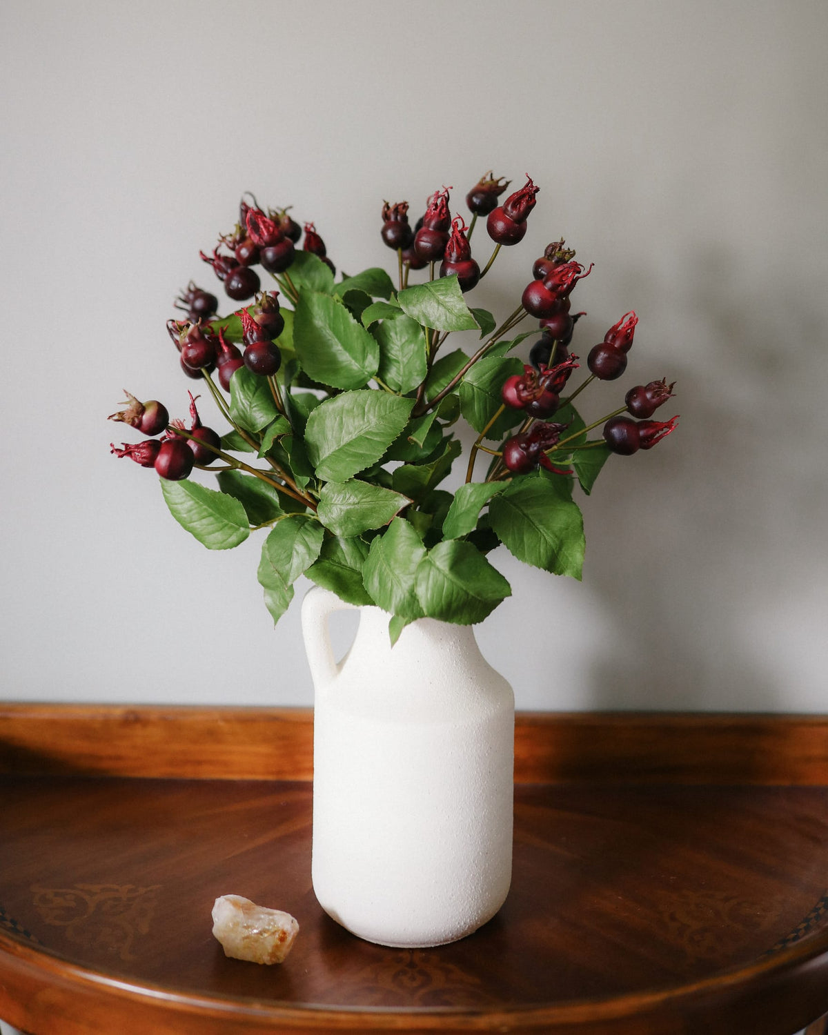 A white ceramic pitcher vase filled with artificial rose hip, displayed on a dark wooden table against a neutral gray wall.