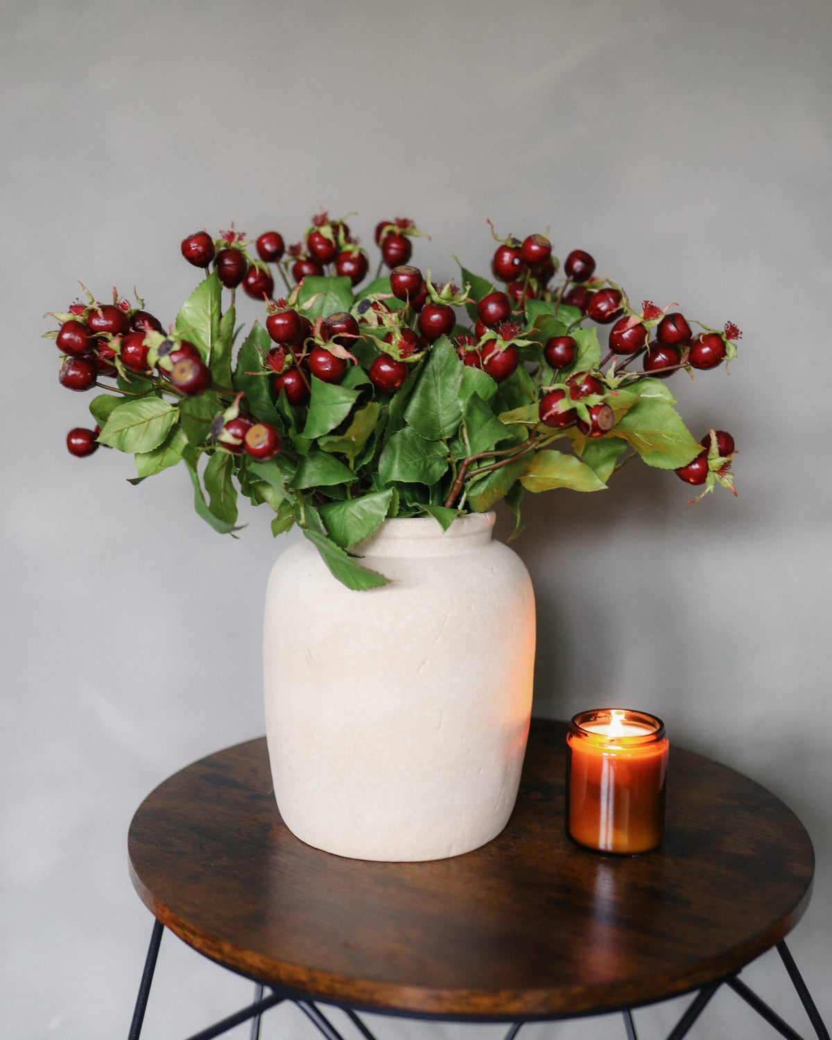 A bouquet of artificial rose hips arranged in a matte off-white ceramic vase, placed on a round dark wood side table next to a lit amber candle against a neutral gray wall.