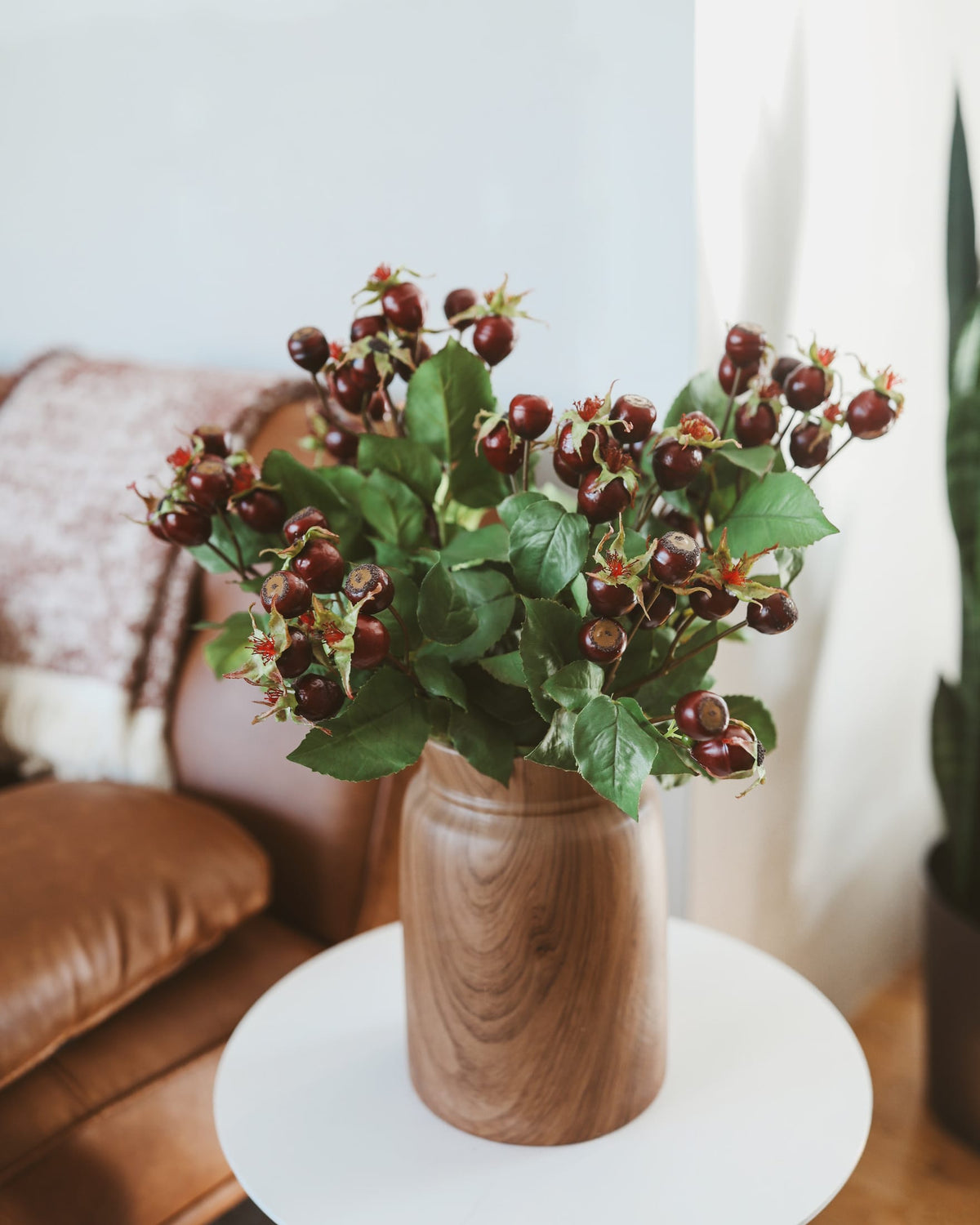 A bouquet of artificial dark rose hips with green leaves arranged in a round wooden vase on a white side table, set in a cozy living room environment.