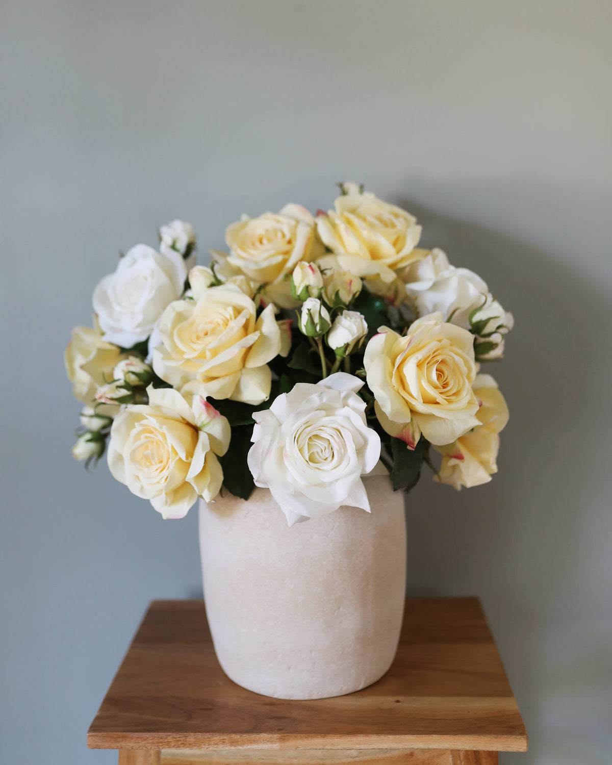 A bouquet of artificial pale yellow and white roses with green foliage arranged in a textured off-white ceramic pot, placed on a wooden surface against a soft gray wall.