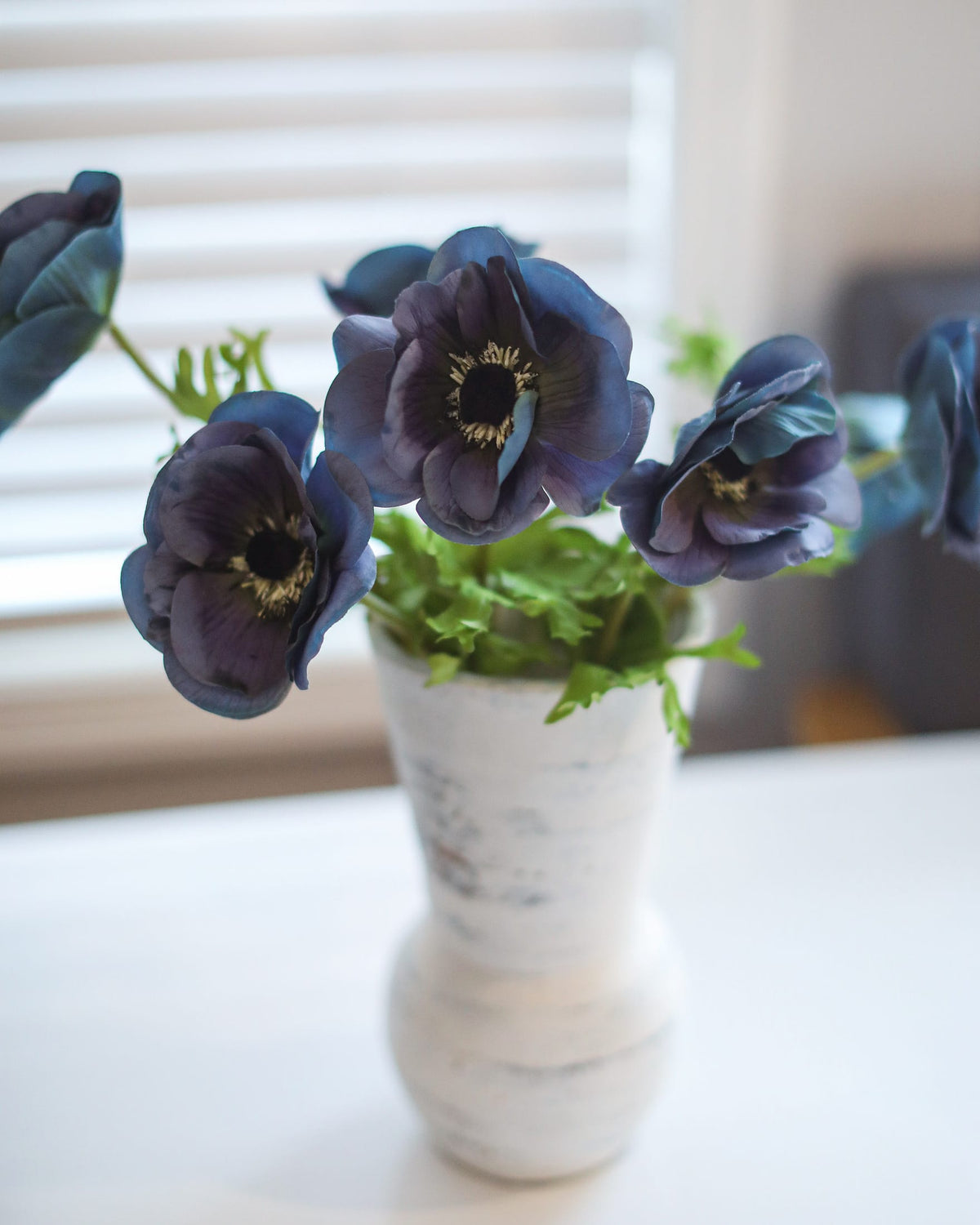 Dark blue faux anemone flowers with green leaves arranged in a white ceramic vase on a white table in front of a softly blurred window with blinds.
