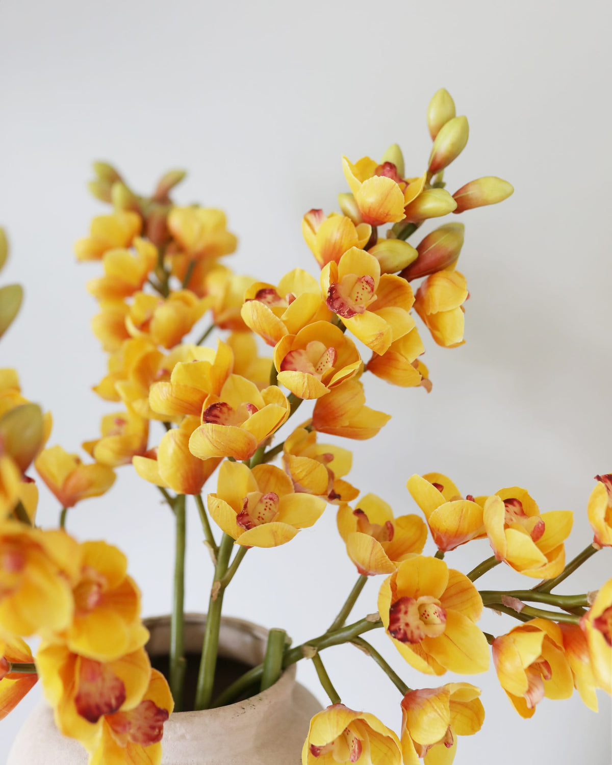 Yellow and orange faux orchid flowers with reddish-purple centers, arranged in a light beige ceramic vase against a soft off-white background.