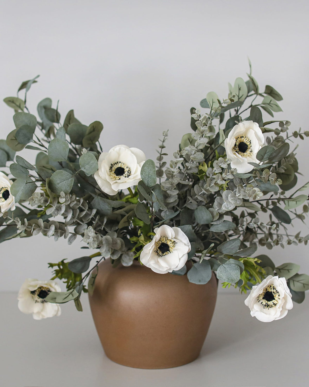 An artificial floral arrangement featuring white faux anemone flowers with dark centers and eucalyptus leaves, presented in a matte brown ceramic pot against a neutral light gray background.