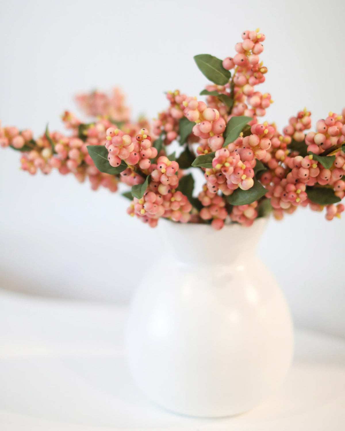 Pink berry stems with green leaves arranged in a small white ceramic vase against a soft white background.