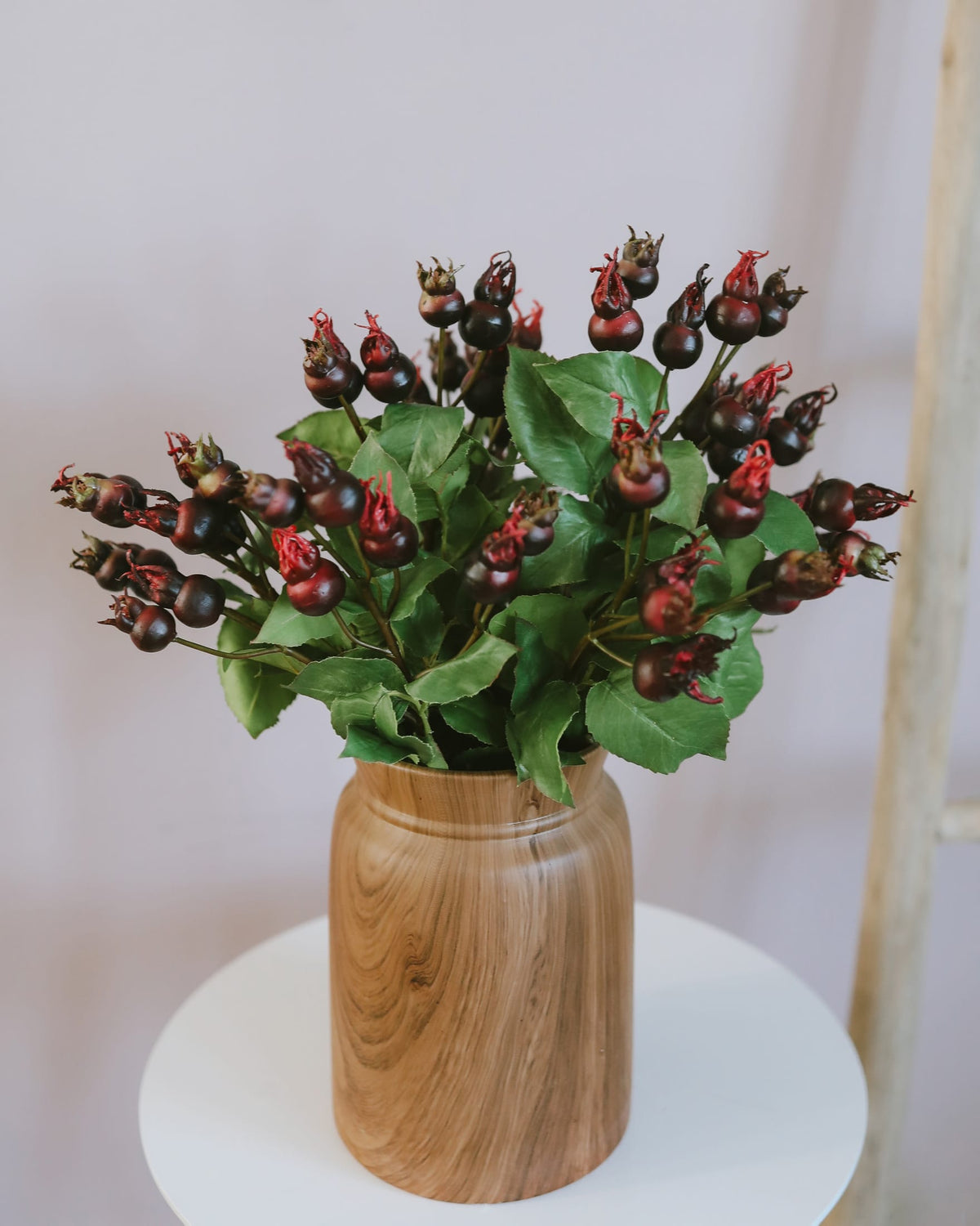 A bouquet of artificial rose hips arranged in a light brown wooden vase, displayed on a white round table against a soft neutral background.