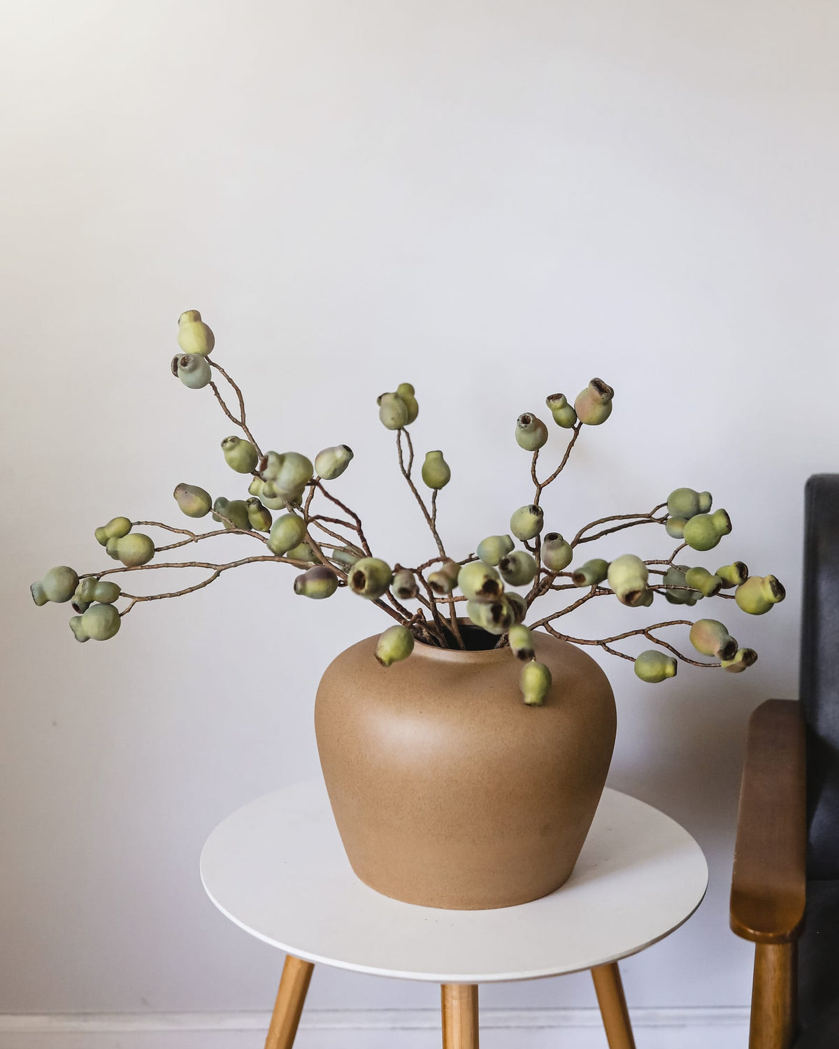 A matte tan ceramic vase filled with faux Eucalyptus Pods, displayed on a small white side table against a plain light wall.