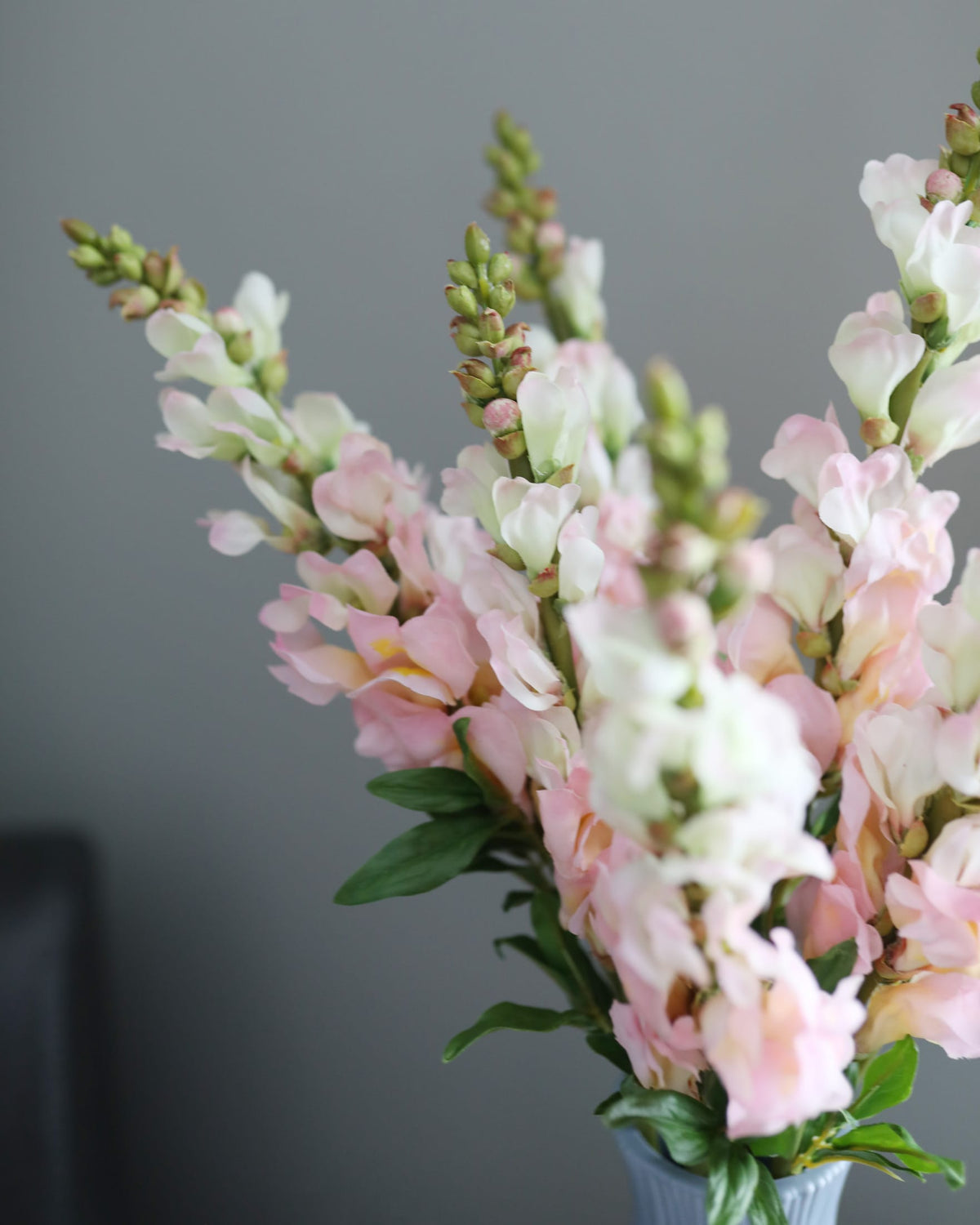 A bouquet of faux pink and white snapdragons with green leaves in a light blue vase against a neutral gray background.