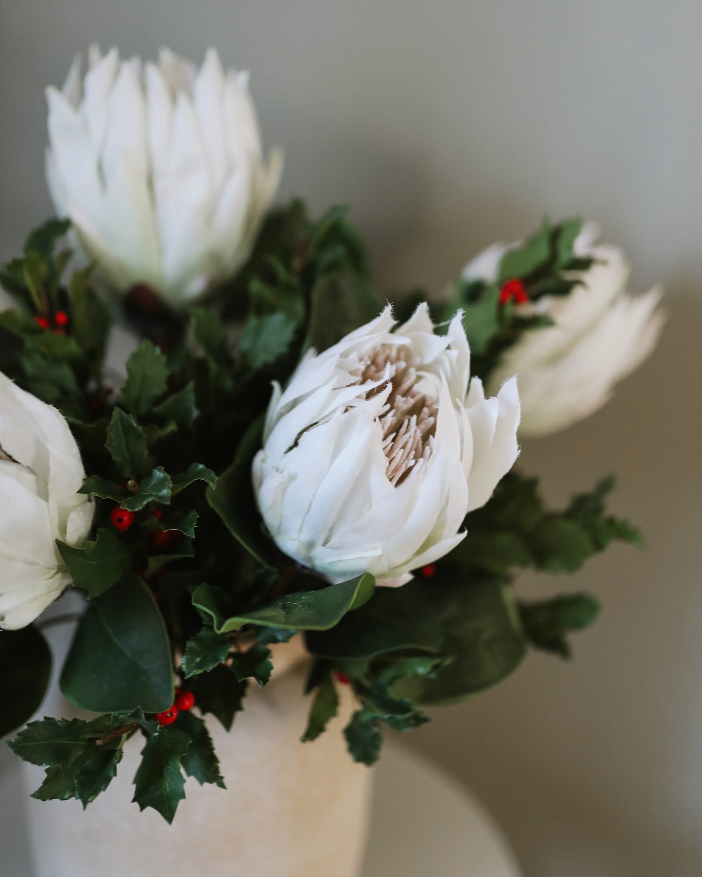 A close-up of a faux white protea flower arrangement with green holly leaves and small red berries in a light-colored vase against a soft neutral background.