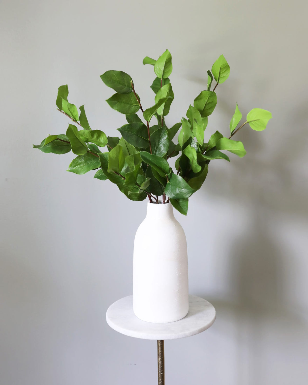 Green artificial salal foliage arranged in a matte white ceramic bottle-shaped vase, displayed on a round white marble side table with a metal stand against a plain light gray wall.