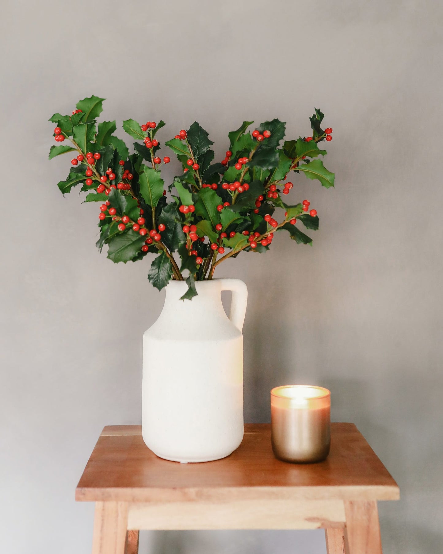 A white ceramic pitcher vase filled with green holly leaves and red berries, placed on a wooden table next to a lit gold candle against a neutral gray wall.